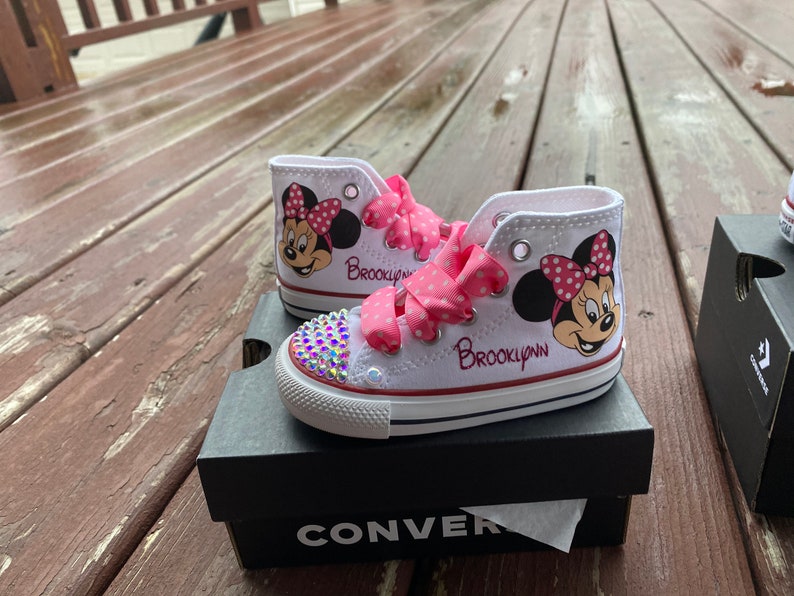 May include: White high-top sneakers with a pink polka dot ribbon, a Minnie Mouse design, and the name "Brooklyn" written on the side. The sneakers are sitting on a black Converse shoebox.