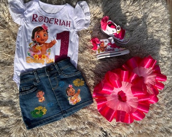 May include: A white onesie with a pink number one and the name "Roderiah" printed on it. The onesie features a cartoon image of Moana from the Disney movie Moana. A pair of white sneakers with a pink bow and a Moana design are also pictured. A denim skirt with Moana patches and a pink tulle tutu are also included.