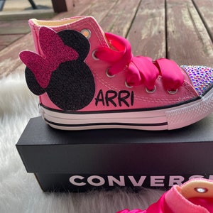 May include: Pink high-top Converse sneakers with a black Minnie Mouse appliqu&eacute; and rhinestones on the toe. The shoes are personalized with the name "ARRI" in black lettering.