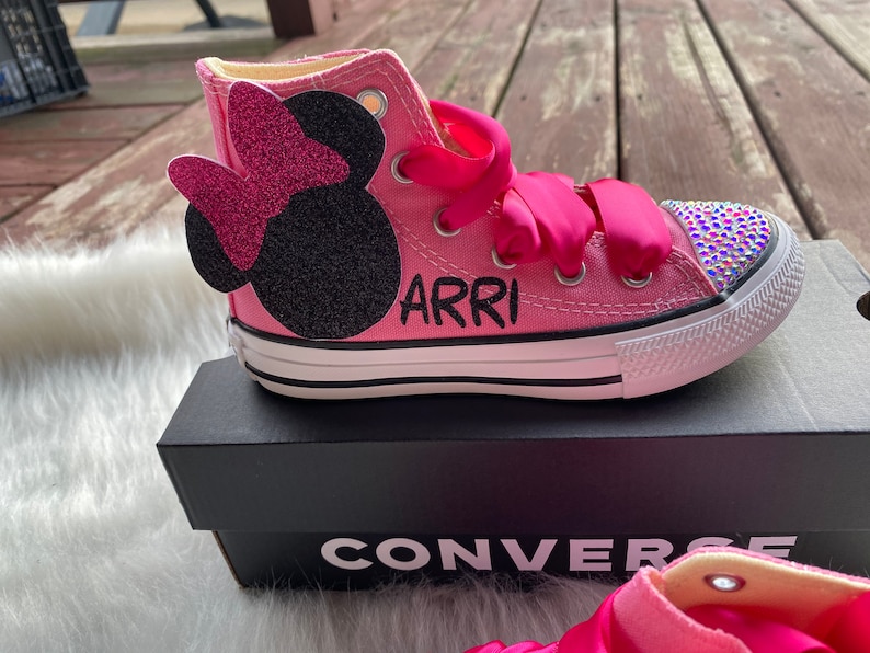May include: Pink high-top Converse sneakers with a black Minnie Mouse appliqu&eacute; and rhinestones on the toe. The shoes are personalized with the name "ARRI" in black lettering.