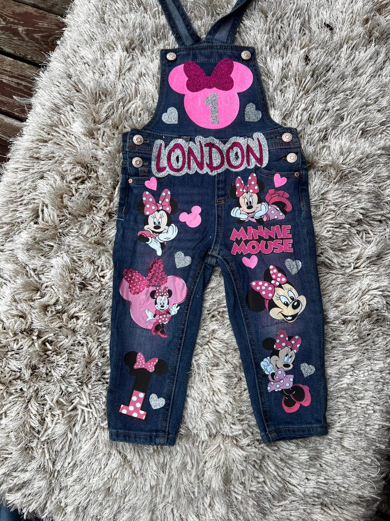K&ouml;nnte beinhalten: Dunkelblaue Jeans-Latzhose mit Minnie-Maus-Motiven. Der Name "LONDON" ist in silbernem Glitzer geschrieben. Die Latzhose hat eine rosa Schleife, die Zahl "1" und verschiedene Minnie-Maus-Bilder und Herzen.