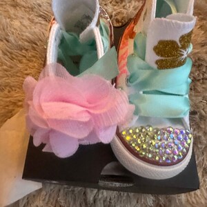 May include: A pair of white baby shoes with pink and blue ribbons, a pink flower, and sparkly rhinestones. The shoes have a gold glitter design on the side.