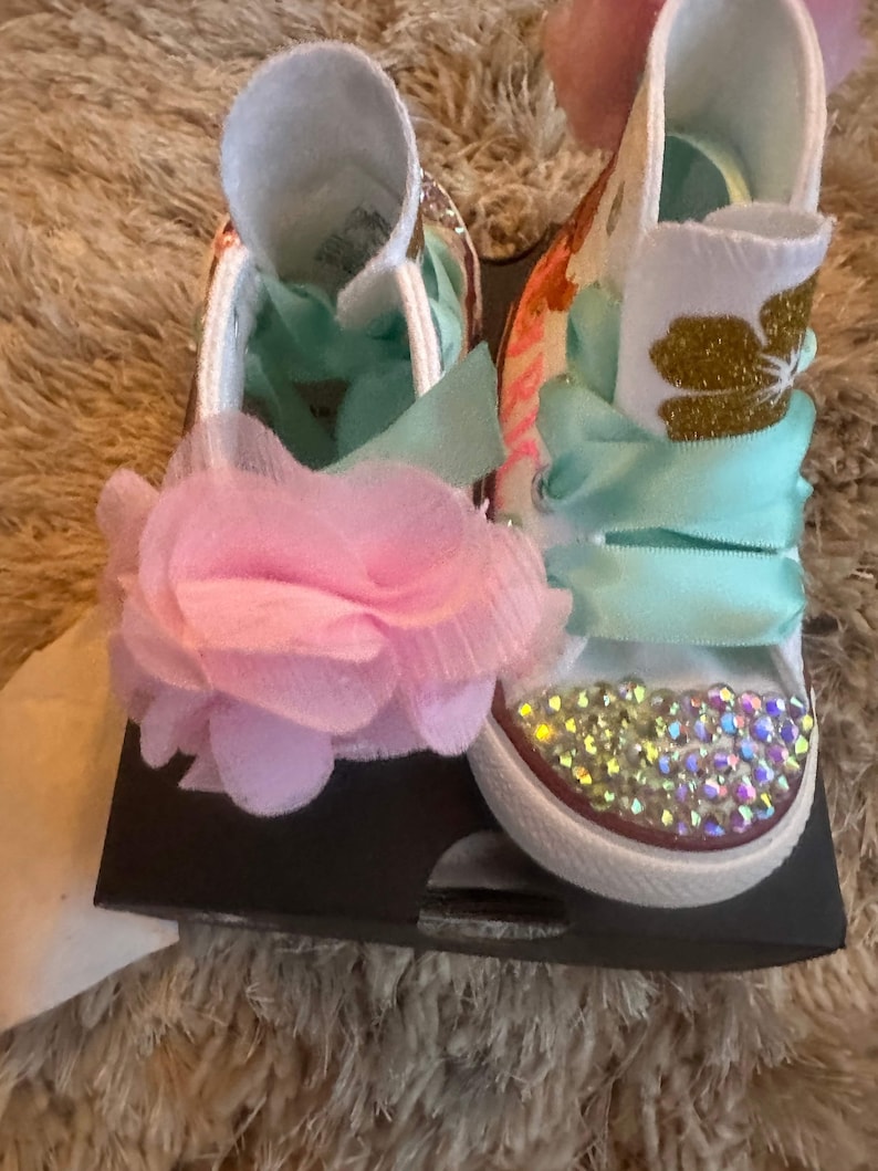 May include: A pair of white baby shoes with pink and blue ribbons, a pink flower, and sparkly rhinestones. The shoes have a gold glitter design on the side.