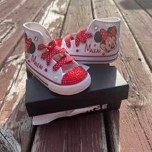 Peut inclure: Baskets montantes blanches avec des d&eacute;tails rouges, repr&eacute;sentant Minnie Mouse et le nom "Maleah". Les bouts des chaussures sont orn&eacute;s de strass rouges, et les lacets sont rouges &agrave; pois blancs. Les baskets sont pr&eacute;sent&eacute;es sur une bo&icirc;te noire.