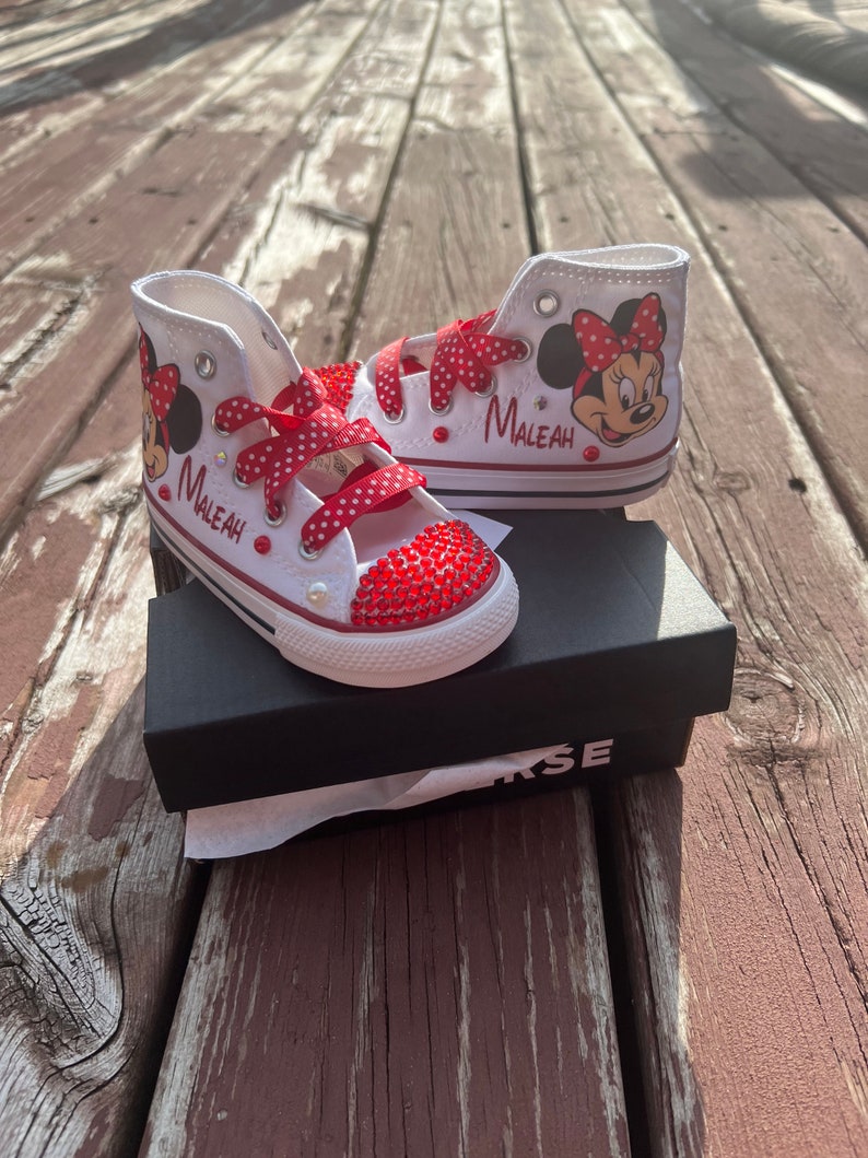 Peut inclure: Baskets montantes blanches avec des d&eacute;tails rouges, repr&eacute;sentant Minnie Mouse et le nom "Maleah". Les bouts des chaussures sont orn&eacute;s de strass rouges, et les lacets sont rouges &agrave; pois blancs. Les baskets sont pr&eacute;sent&eacute;es sur une bo&icirc;te noire.