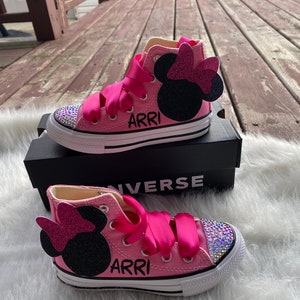 May include: Pink high-top Converse sneakers with a black Minnie Mouse appliqu&eacute; and pink bow. The shoes are decorated with rhinestones and have the name "ARRI" written on them.