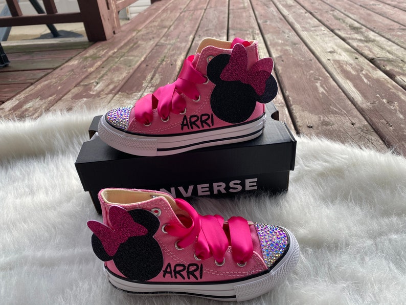 May include: Pink high-top Converse sneakers with a black Minnie Mouse appliqu&eacute; and pink bow. The shoes are decorated with rhinestones and have the name "ARRI" written on them.