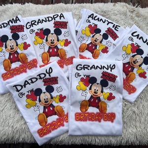 Pu&ograve; includere: Magliette bianche con grafiche di Topolino e testi per i membri della famiglia: Nonna, Nonno, Zia, Zio, Pap&agrave; e Nonna. Ogni maglietta ha il testo "Birthday Boy".