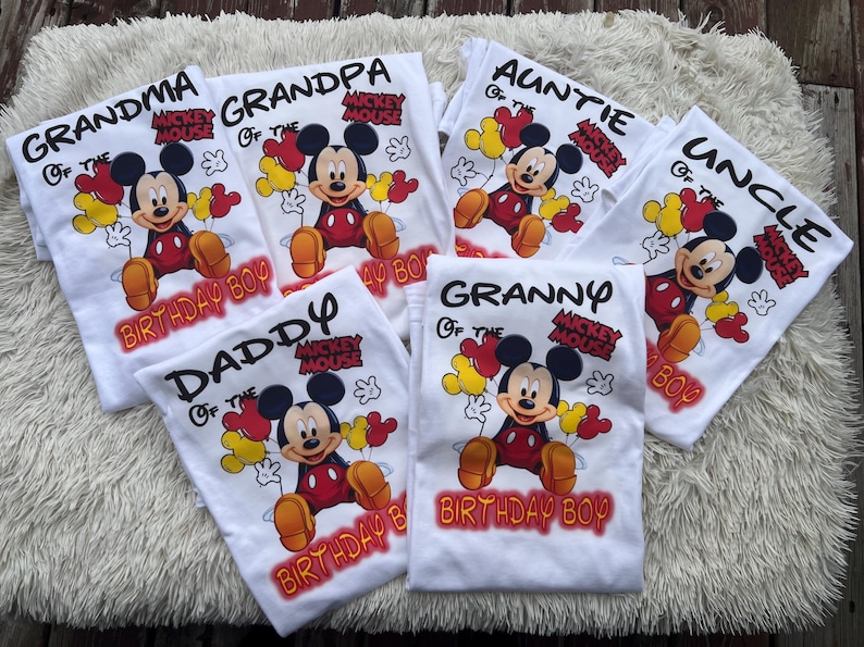 Pu&ograve; includere: Magliette bianche con grafiche di Topolino e testi per i membri della famiglia: Nonna, Nonno, Zia, Zio, Pap&agrave; e Nonna. Ogni maglietta ha il testo "Birthday Boy".