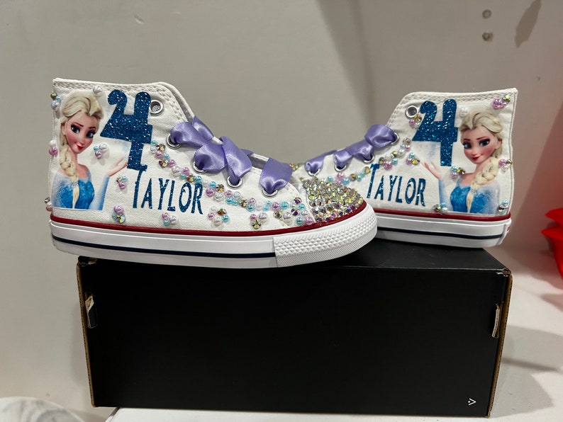 Puede incluir: Zapatillas blancas de ca&ntilde;a alta con una cinta morada, purpurina azul y una imagen de Elsa de Frozen. Las zapatillas est&aacute;n decoradas con pedrer&iacute;a y dicen "Taylor" y "4".