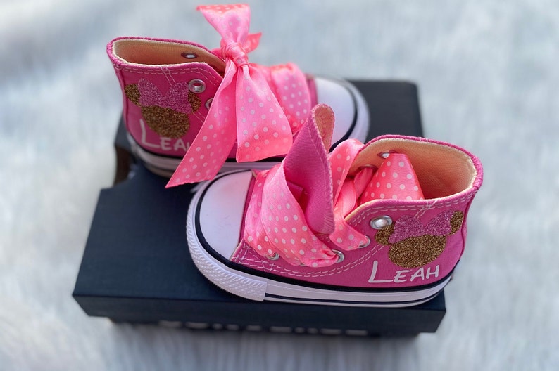 Puede incluir: Zapatillas altas de color rosa con cordones de cinta de lunares blancos y un dise&ntilde;o de Minnie Mouse con purpurina dorada. Las zapatillas est&aacute;n personalizadas con el nombre "Leah".