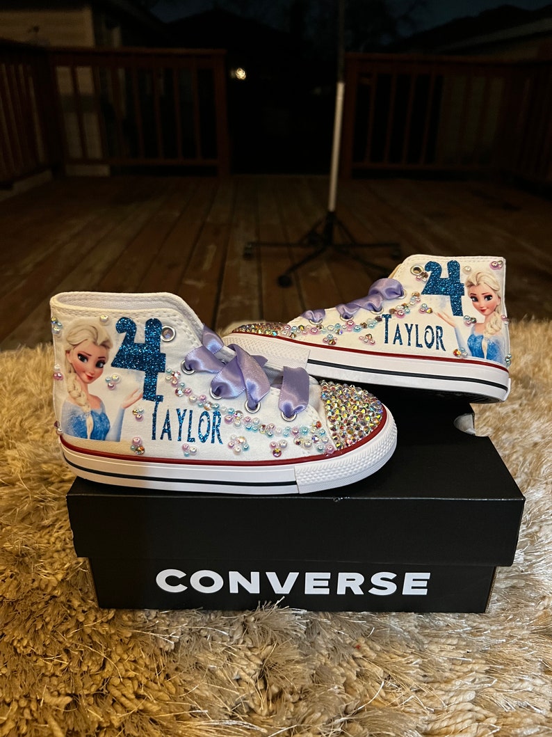 Puede incluir: Zapatillas Converse blancas de ca&ntilde;a alta con una imagen azul y blanca de Elsa de Frozen, un n&uacute;mero "4" azul y el nombre "Taylor" en purpurina azul. Las zapatillas est&aacute;n decoradas con pedrer&iacute;a y tienen una cinta de raso morado.