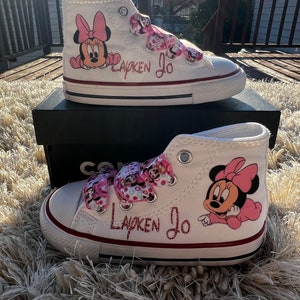 May include: White high-top sneakers with pink Minnie Mouse character designs and pink ribbon laces. The shoes are personalized with the name "Layken Jo" in pink glitter.