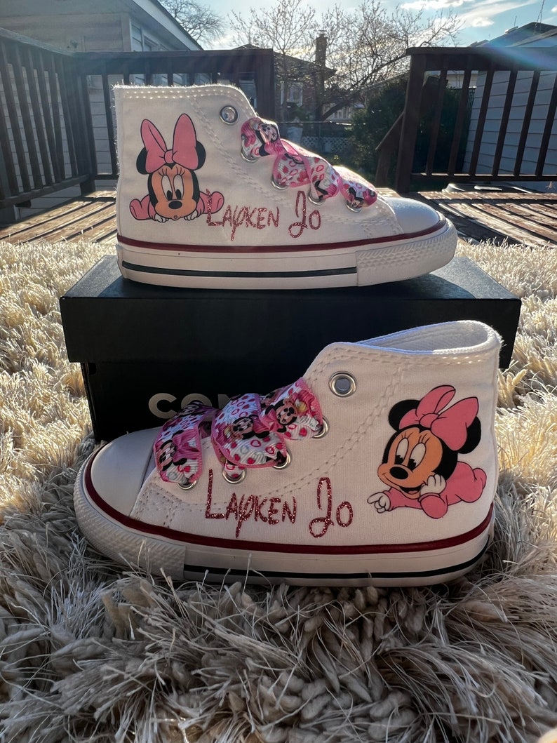 May include: White high-top sneakers with pink Minnie Mouse character designs and pink ribbon laces. The shoes are personalized with the name "Layken Jo" in pink glitter.