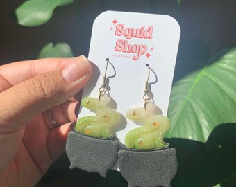 May include: A pair of dangle earrings featuring a matte black cauldron base with a swirling, translucent light green smoke effect rising from the top. The smoke includes small gold star accents. The earrings are displayed on a white card with the text Squid Shop Creations printed in red.