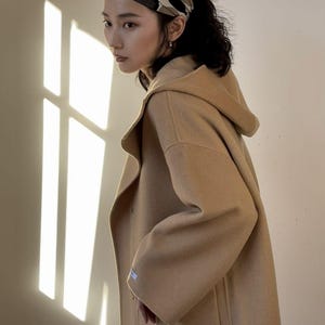 May include: A tan hooded coat with a large pocket and a matching headband with a black and brown pattern. The coat is a neutral color and appears to be made of a soft material. The image is lit by sunlight.