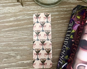 May include: A rectangular bookmark with a repeating pattern of martini glasses and pink bows on a light pink background. The bookmark is next to a book with a floral cover and a candle.