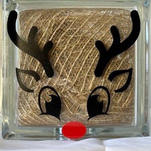 May include: A glass block with a reindeer face design. The reindeer has black antlers, eyes, and a nose. The nose is red and the background is a brown hessian material.