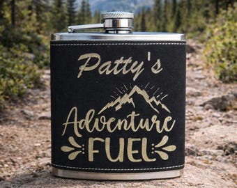 Könnte beinhalten: Eine schwarze Flasche mit silbernem Deckel und schwarzem Lederbezug. Auf der Flasche steht in goldener Schrift "Patty's Adventure FUEL". Der Hintergrund zeigt eine Bergkette und Bäume.