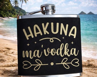 Könnte beinhalten: Eine schwarze Flachmann mit silbernem Verschluss und Scharnier, mit goldfarbenem Schriftzug "HAKUNA ma' vodka". Der Flachmann steht an einem Sandstrand mit tropischem Ozean-Hintergrund.