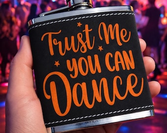 Könnte beinhalten: Eine schwarze Flachmann mit silbernem Deckel und Zierleiste. Auf dem Flachmann steht in orangefarbener Schrift "Trust Me You Can Dance". Der Flachmann wird in einer Hand gehalten, mit einem verschwommenen Hintergrund einer Tanzfläche.