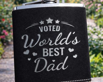 Könnte beinhalten: Eine schwarze Flachmann mit silbernem Verschluss und Zierleiste. Der Flachmann trägt den silbernen Schriftzug "Voted World's Best Dad". Der Hintergrund ist eine verschwommene Außenszene mit Grün.