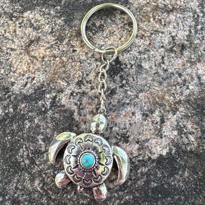 May include: A silver-toned metal keychain featuring a turtle-shaped charm. The turtle shell is decorated with etched patterns and a central circular turquoise stone. The charm is attached to a standard silver-colored metal split ring by a short chain.