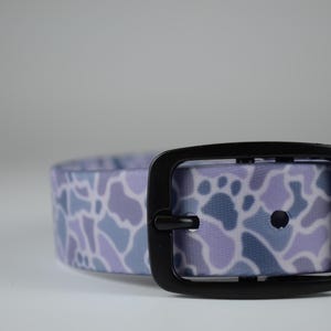 May include: A pet collar featuring a light purple, lavender, and slate blue camouflage pattern with a matte black metal buckle and a matching black metal keeper loop. The strap has a smooth, flexible texture with pre-punched holes for adjustment.