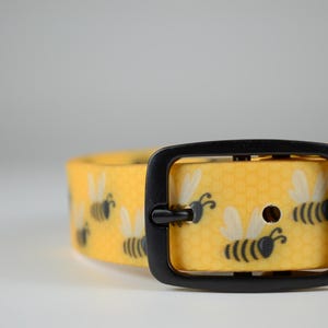 May include: A yellow collar featuring a repeating pattern of black and white bees over a subtle hexagonal honeycomb background. The collar includes a matte black metal buckle and a matching black metal loop for secure fastening and adjustment.