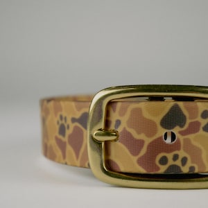 May include: A close-up of a dog collar with a camouflage pattern in shades of tan, brown, and black. The collar features a gold-colored metal buckle. The pattern includes paw prints.