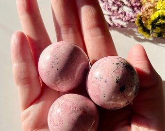 May include: Three pink and white polished spheres of pink gemstone, likely rhodonite, in a hand.