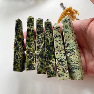 May include: Seven green and black speckled stone points, also known as green moss agate, are arranged in a row. The points are smooth and polished.
