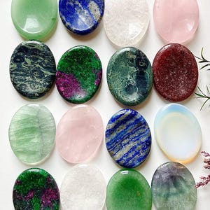 May include: Sixteen polished, oval gemstones in a variety of colours. The gemstones include green, blue, white, pink, and other colours. The stones are arranged in four rows of four, showcasing their unique textures and hues.