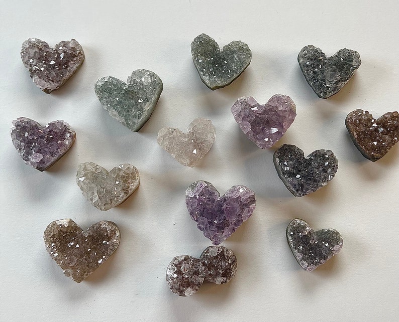 Puede incluir: Una colecci&oacute;n de catorce cristales de amatista en forma de coraz&oacute;n en varios tonos de morado, gris y blanco. Los cristales est&aacute;n dispuestos sobre una superficie blanca.
