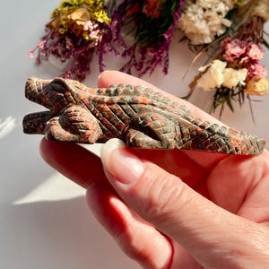 May include: A small, hand-carved figurine of an alligator crafted from a mottled, reddish-brown and dark green stone. The sculpture features detailed, textured scales along its back, a defined snout, and a long, tapering tail in a horizontal pose.