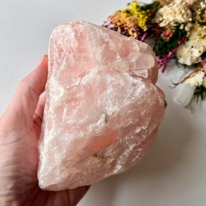 May include: A large, raw chunk of rose quartz crystal with a translucent, pale pink hue. The stone features a jagged, uneven surface with natural crystalline fractures and varying levels of opacity throughout its irregular, multi-faceted structure.