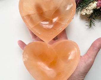 May include: Two heart-shaped bowls made of orange selenite crystal. The bowls are smooth and polished, with a natural, slightly translucent appearance.
