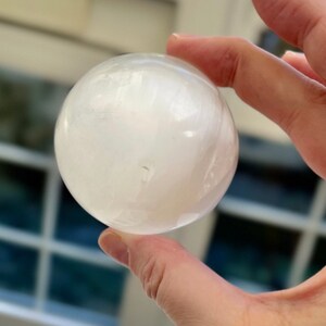 May include: A translucent, white sphere, possibly a crystal ball or decorative object. The sphere is held in a hand, with the background slightly blurred. The surface of the sphere appears smooth and reflective, with subtle internal patterns.