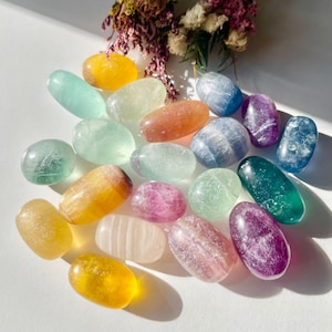 May include: A selection of polished gemstones in shades of yellow, green, blue, purple, and pink. The oval-shaped stones are translucent, displaying natural colour and texture variations. Dried flowers are visible in the background.