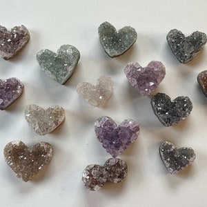 Puede incluir: Una colecci&oacute;n de catorce cristales de amatista en forma de coraz&oacute;n en varios tonos de morado, gris y blanco. Los cristales est&aacute;n dispuestos sobre una superficie blanca.