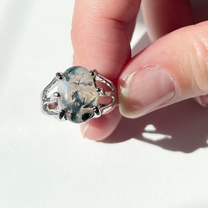 May include: A silver ring with a large oval-shaped moss agate stone. The stone has a white, gray, and green color pattern.