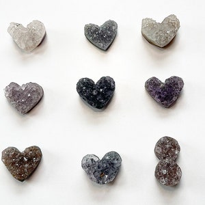 Puede incluir: Nueve racimos de cristal de amatista en forma de coraz&oacute;n en varios tonos de morado, gris y blanco. Los cristales est&aacute;n dispuestos en una cuadr&iacute;cula sobre un fondo blanco.