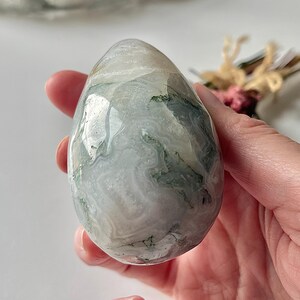 May include: A polished, egg-shaped moss agate stone featuring a smooth, glossy surface with translucent white, gray, and milky tones, accented by intricate, dark green, fern-like dendritic inclusions throughout the mineral structure.