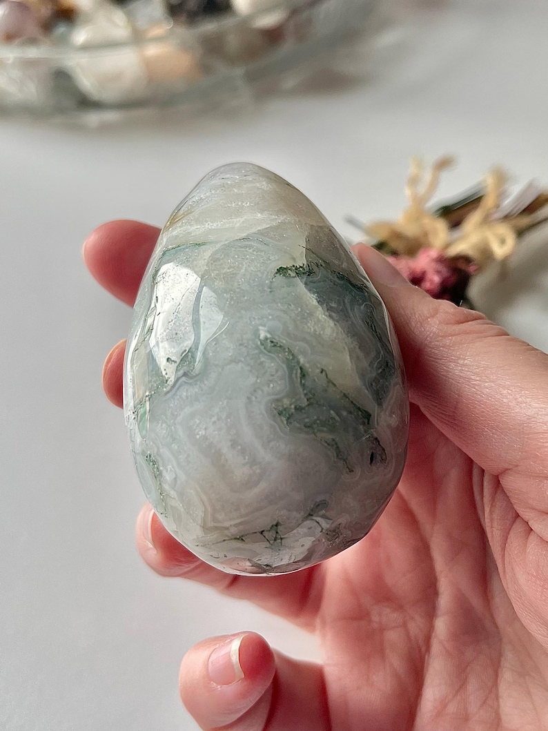 May include: A polished, egg-shaped moss agate stone featuring a smooth, glossy surface with translucent white, gray, and milky tones, accented by intricate, dark green, fern-like dendritic inclusions throughout the mineral structure.