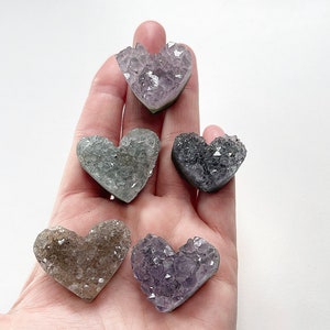 Puede incluir: Cinco cristales de amatista en forma de coraz&oacute;n en varios tonos de morado, verde y marr&oacute;n. Los cristales est&aacute;n agrupados en una mano.