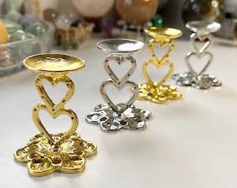 May include: Three decorative stands made of metal with a heart design. The stands are gold, silver, and gold. The stands are on a white surface.