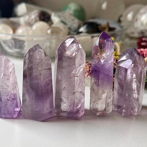 May include: Five amethyst crystal points of varying sizes stand upright. The crystals are a translucent purple colour with visible internal structures. The background includes other crystals and decorative elements.