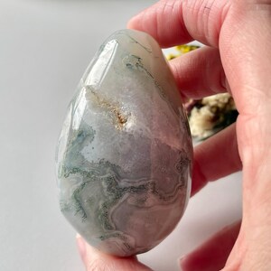 May include: A polished, egg-shaped stone with a translucent appearance. The stone features a mix of white, gray, and green patterns, creating a natural, organic aesthetic. The smooth surface reflects light, highlighting the intricate veining within the stone.