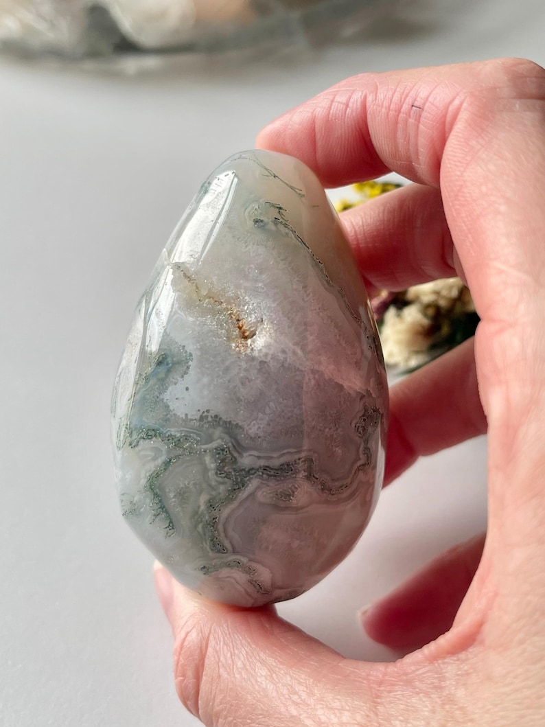 May include: A polished, egg-shaped stone with a translucent appearance. The stone features a mix of white, gray, and green patterns, creating a natural, organic aesthetic. The smooth surface reflects light, highlighting the intricate veining within the stone.