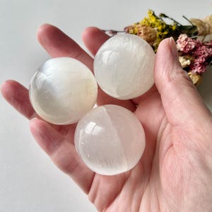 May include: Three white selenite spheres, each with a smooth, polished surface. The spheres are held in an open hand, with a small bouquet of dried flowers in the background. The spheres are a milky white color.
